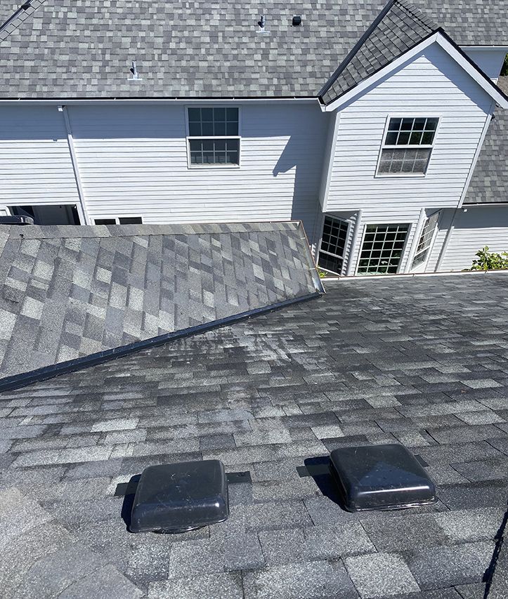 A roof with two vents on it and a white house in the background.