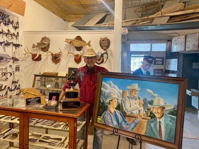 Man in cowboy hat holding a framed painting inside a museum, displaying artifacts and taxidermy.