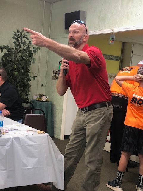 Man in red shirt points with a microphone, speaking in a room with a table and another person wearing orange.