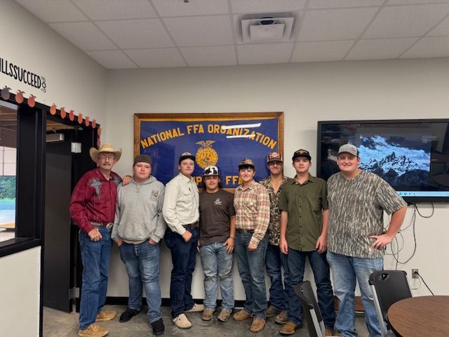 Group of FFA members posing in front of a National FFA Organization sign. They are in a room and wearing hats.