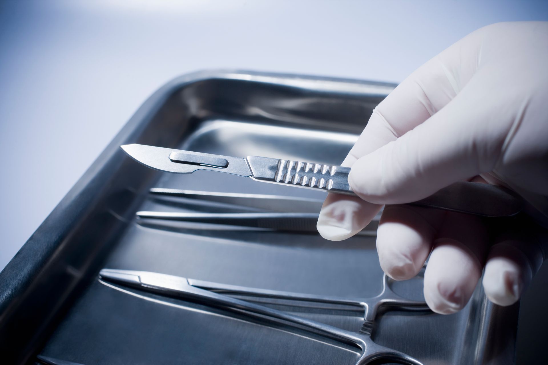 Gloved hand holding a scalpel over a metal surgical tray with instruments