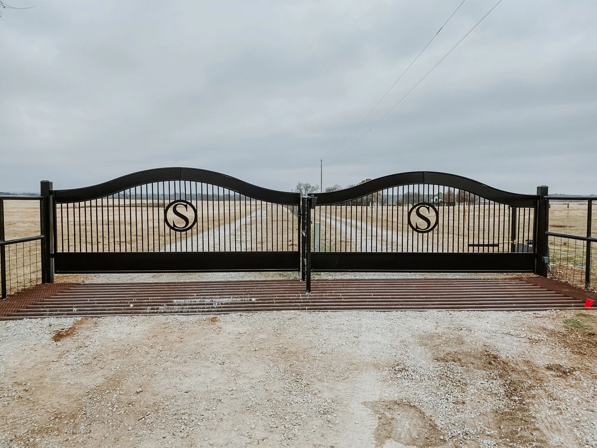Black metal gate with curved top on a gravel driveway under a cloudy sky.