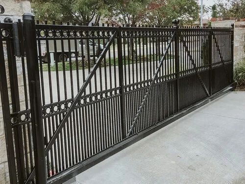 Black wrought-iron sliding driveway gate beside a concrete sidewalk in front of a house