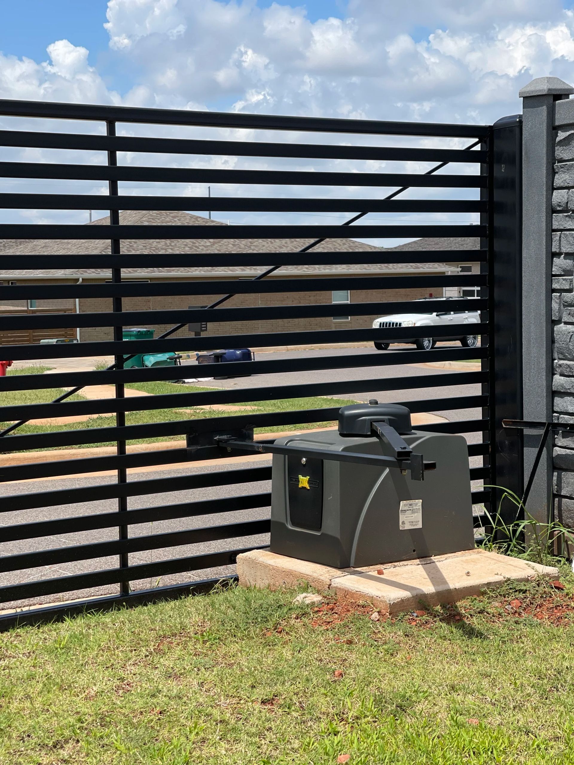 Black slatted fence with a gray utility box on a concrete pad beside a grassy yard, under a cloudy sky