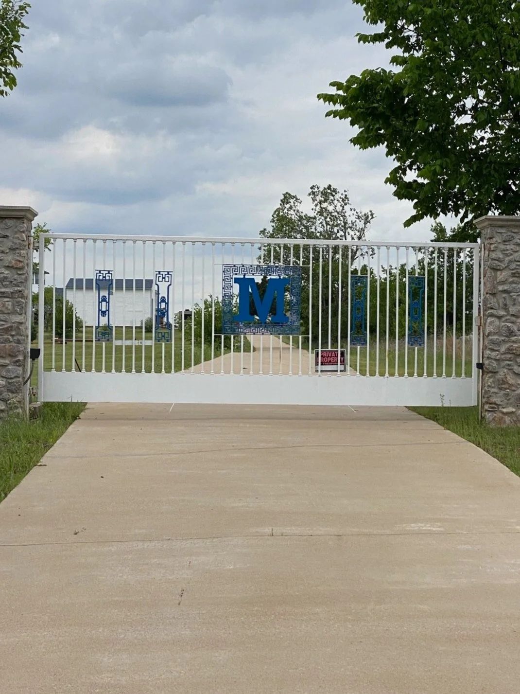 White gate with blue banners and a large blue “M” at the end of a driveway, under a cloudy sky