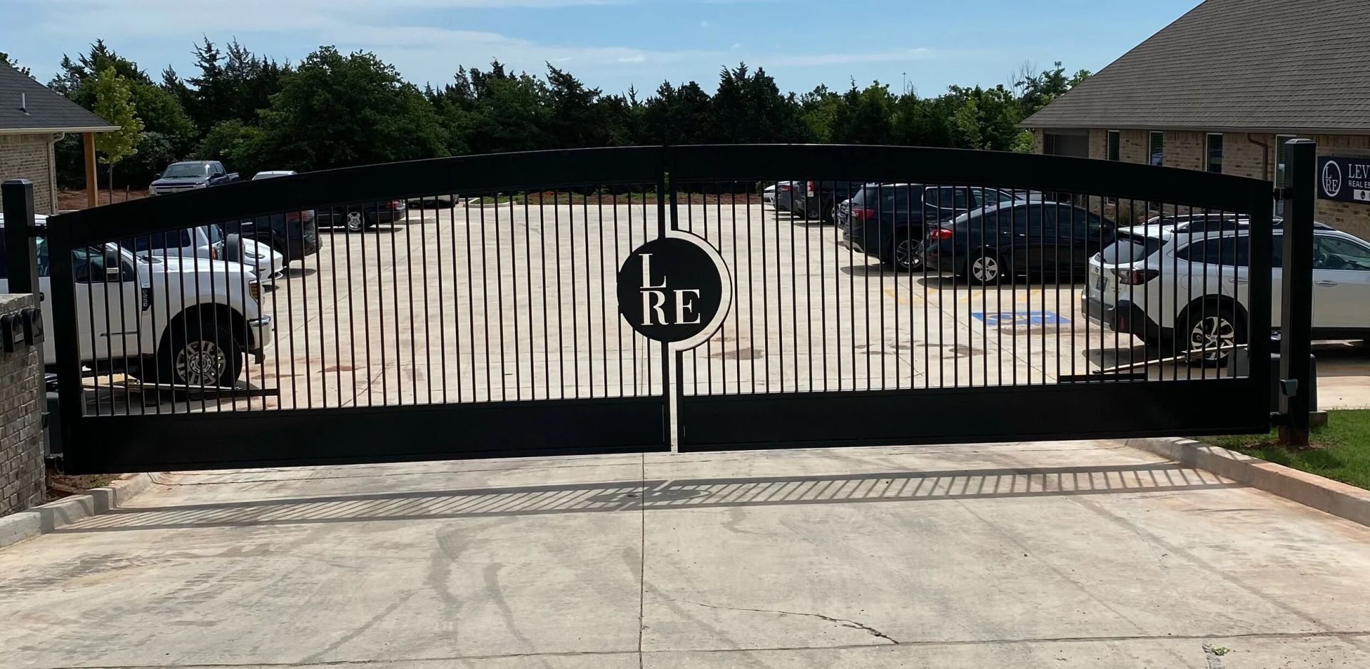 Black metal gate with round emblem at driveway entrance, cars parked behind it.