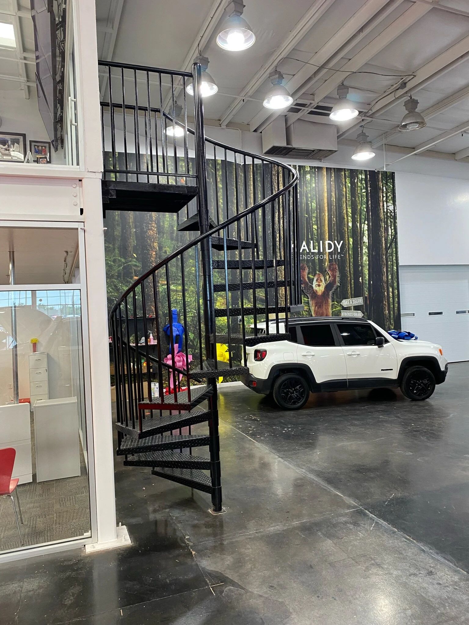 White SUV parked indoors beside a black spiral staircase in an industrial-style showroom.