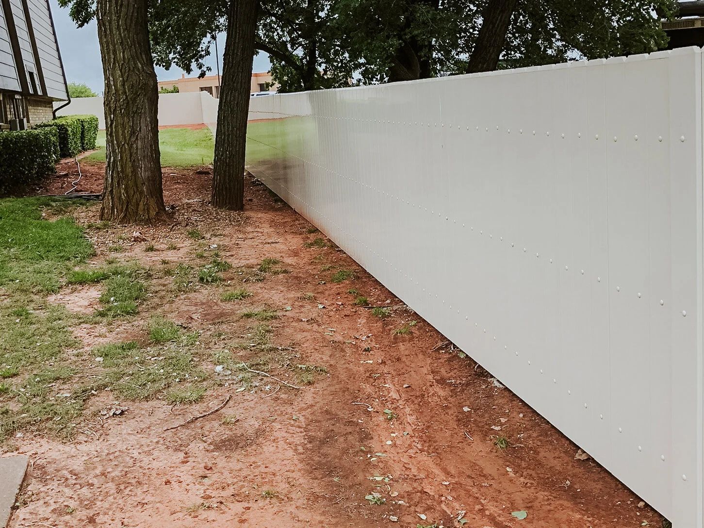 Dirt path beside a long white fence with trees and a house in the background