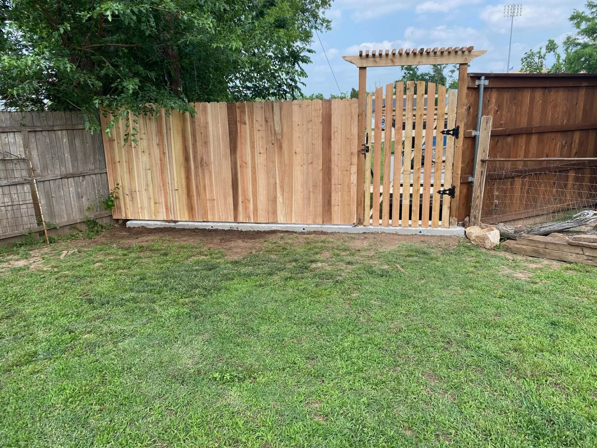 Backyard with green lawn, wooden fence, and a light wood gate under a pergola.