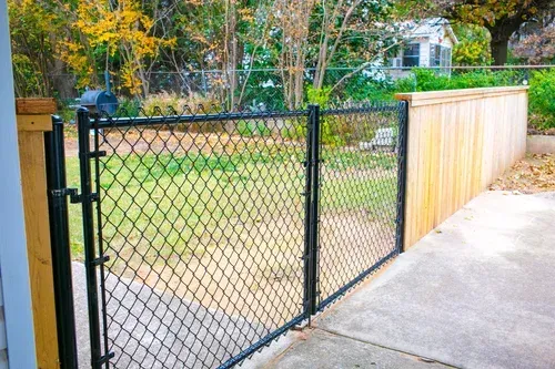 Black chain-link gate and wooden fence beside a concrete driveway in a leafy backyard