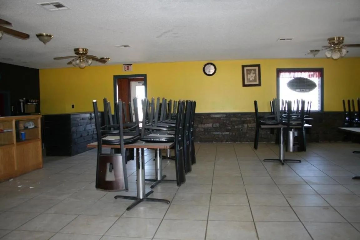 Interior of a restaurant with yellow walls, dark tile wainscoting, a counter, and tables with stacked chairs on tile floor.