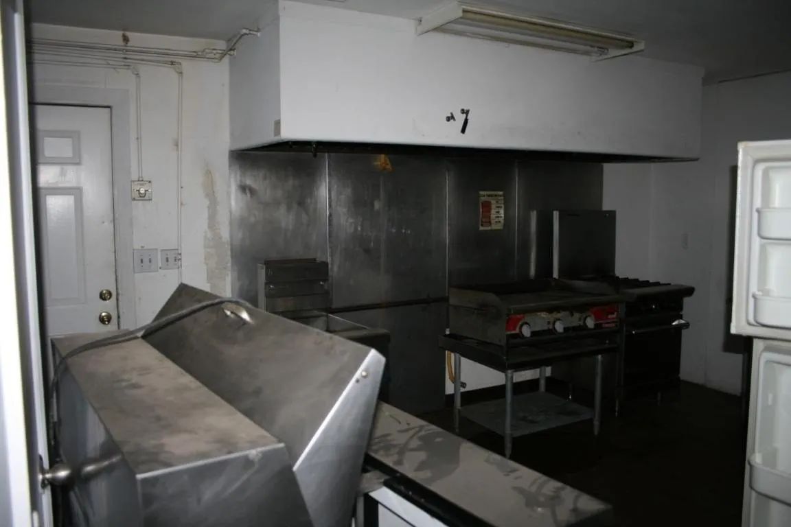 A dim commercial kitchen with stainless steel counters, a vent hood, a stove, a white door, and a refrigerator.
