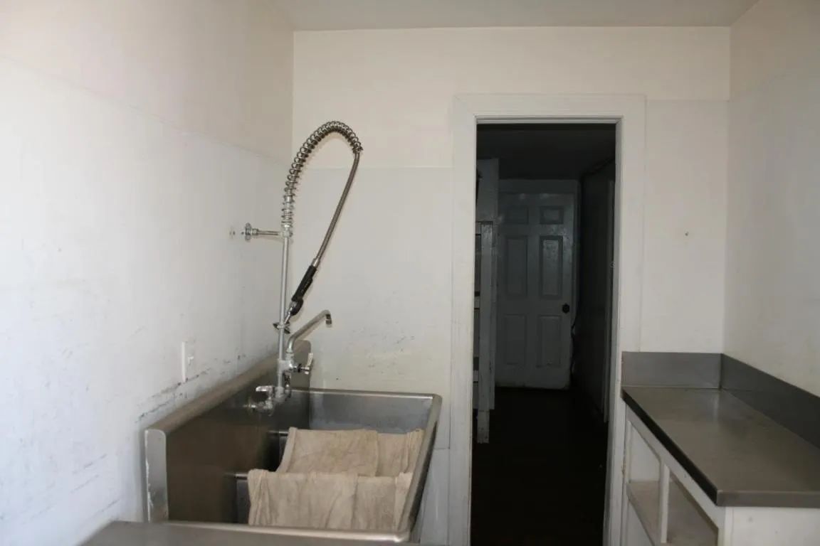 A commercial stainless steel sink with a tall spray faucet sits against a white wall next to a doorway.