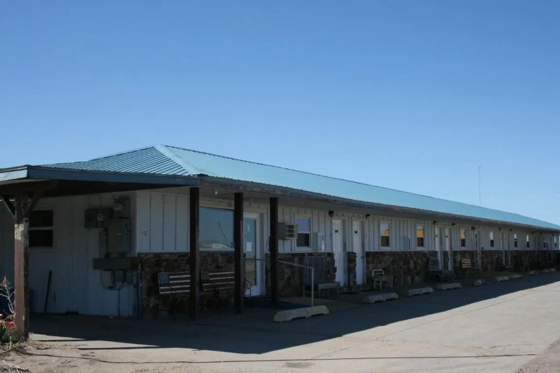 A single-story motel with a light blue metal roof, stone foundation, and multiple doors along an exterior walkway.