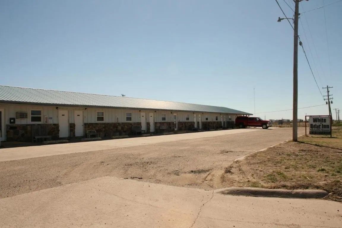 Single-story motel with white siding, stone accents, and a metal roof, featuring a gravel parking lot and roadside sign.