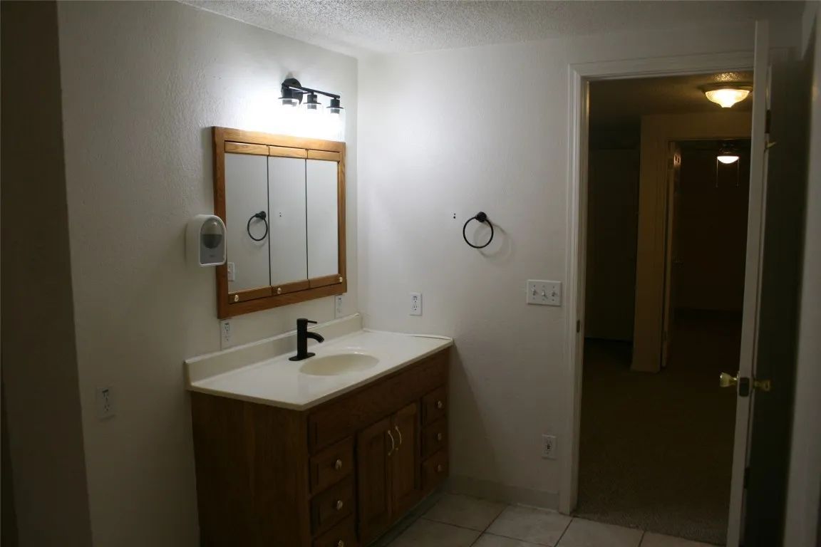 A bathroom vanity with a wood cabinet, white countertop, sink, mirror, and light fixture, with a doorway in the background.