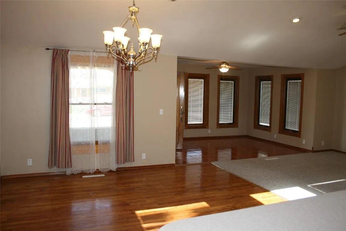 A dining room with hardwood floors, a gold chandelier, and a living area with windows and carpet in the background.
