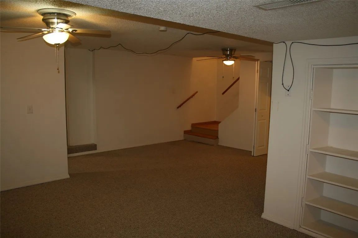 An empty, beige-carpeted basement room with two ceiling fans, white walls, and built-in wall shelving.
