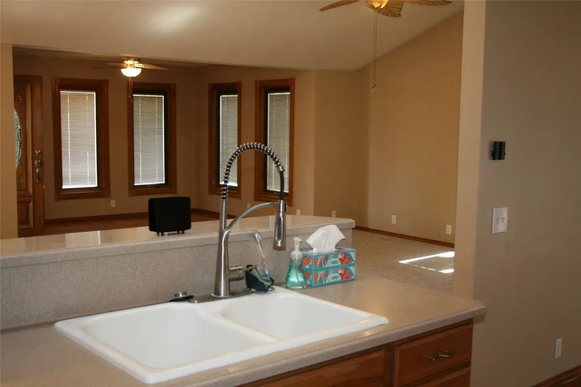 A kitchen sink and faucet in the foreground, with an open-plan living room featuring windows and a ceiling fan behind it.
