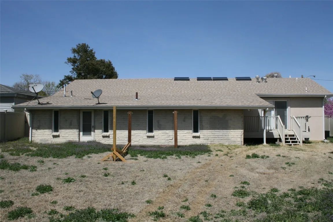 Single-story light stone house with a shingled roof, solar panels, and a dry, sparse lawn under a clear blue sky.