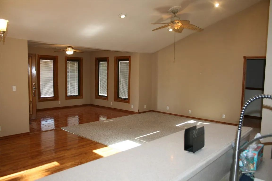 A spacious, neutral-toned living room with hardwood floors, a carpeted area, large windows, and a ceiling fan.