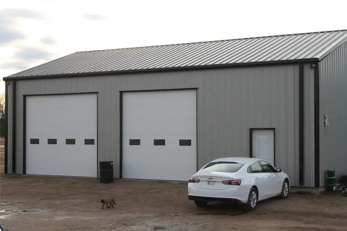 A gray metal workshop with two white garage doors, a side entry door, and a white sedan parked in front on a dirt lot.