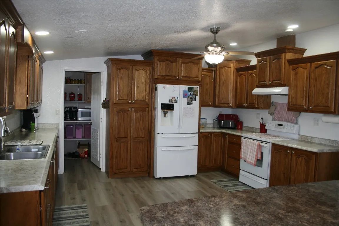 A well-lit kitchen features medium-toned wooden cabinets, white appliances, countertops, and a doorway to a pantry.