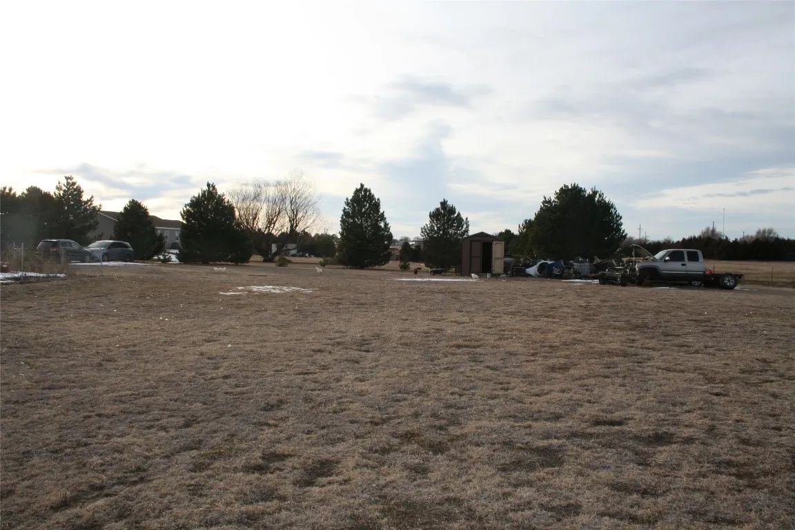 A wide view of a dry, grassy field with several evergreen trees, a small shed, and a truck parked under a cloudy sky.
