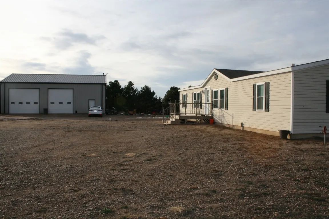 A gravel lot sits between a beige single-story manufactured home and a large metal garage building under a cloudy sky.