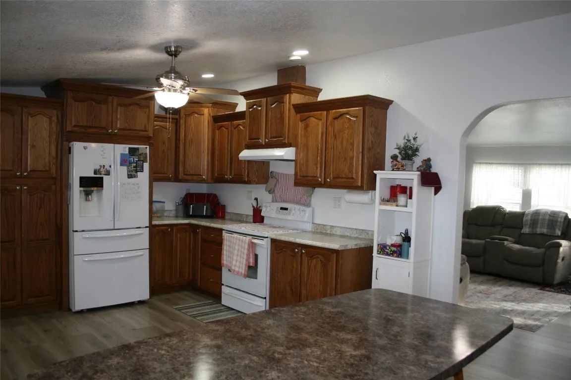 A spacious kitchen with oak cabinets, white appliances, a ceiling fan, and an arched doorway leading to a living area.