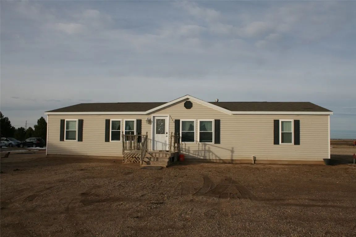 A single-story, beige manufactured home with brown shutters, a central entrance, and a dirt yard under a cloudy sky.