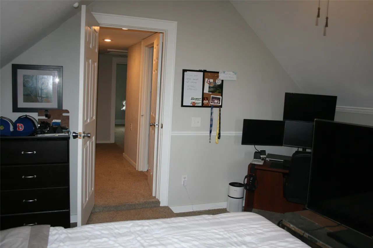 A bedroom with a dresser, an open doorway leading to a hallway, a bulletin board, and a computer desk with multiple monitors.