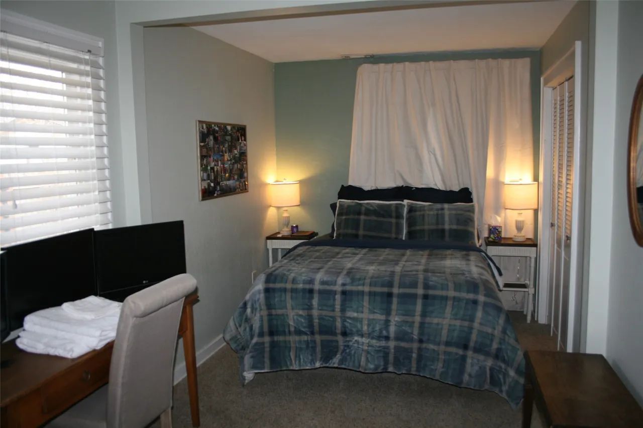 A bedroom with a queen bed featuring a blue plaid comforter, two nightstands, a desk with a chair, and window blinds.
