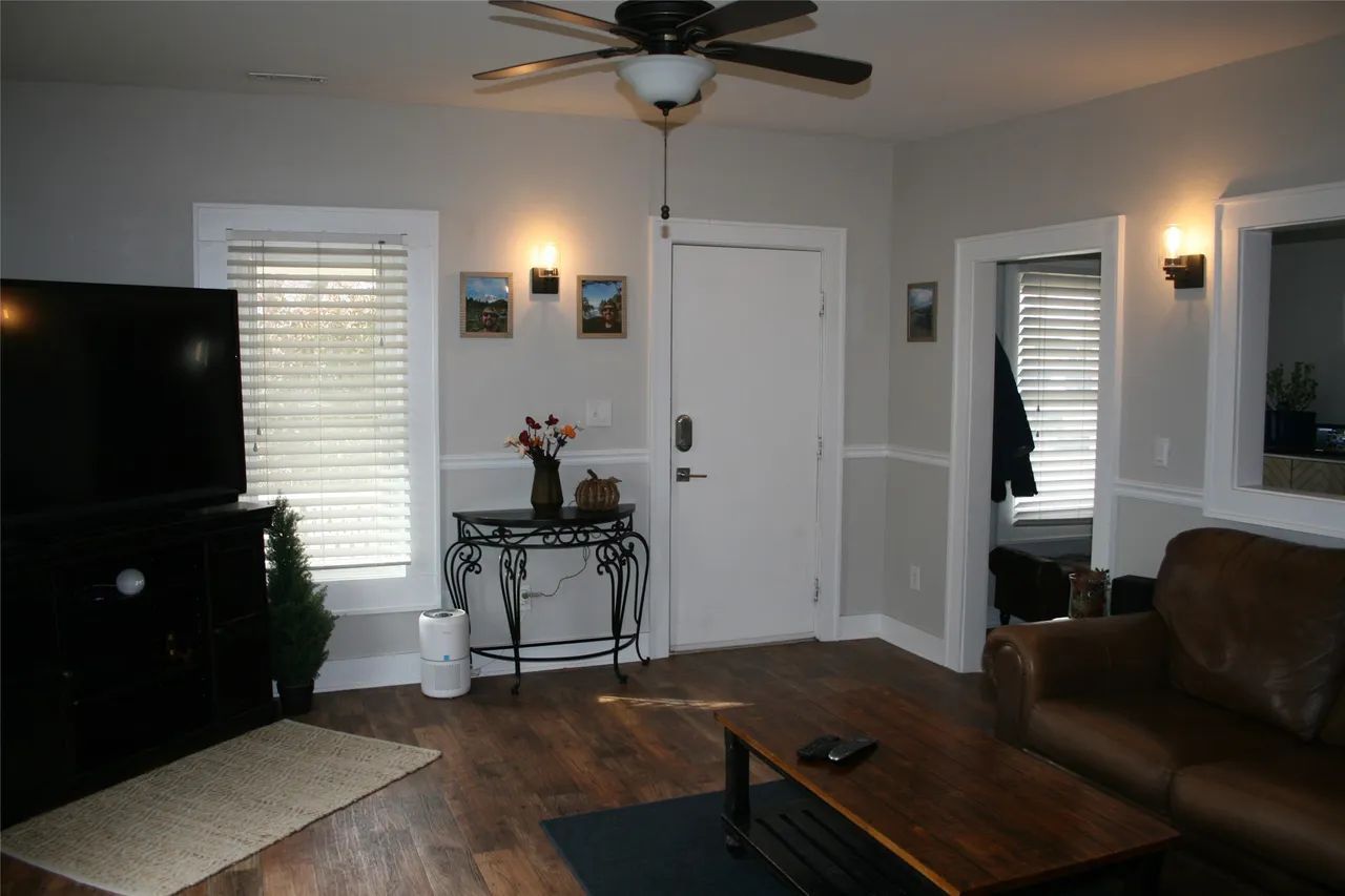 A living room with light gray walls, a ceiling fan, a large TV, a brown sofa, a dark coffee table, and a white front door.