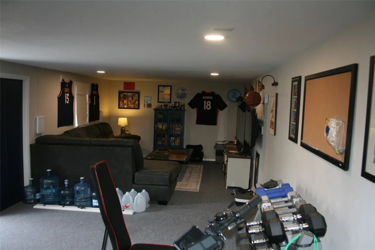 A basement living room features a dark gray sectional sofa, a weight rack with dumbbells, and sports jerseys on the walls.