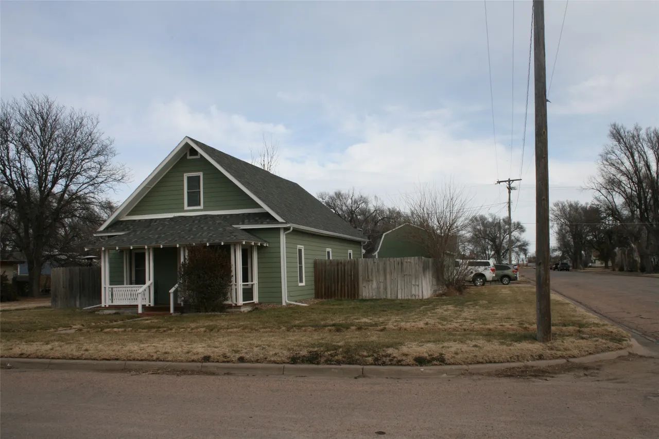 A green, single-story house with a porch sits on a grassy corner lot under a cloudy sky.