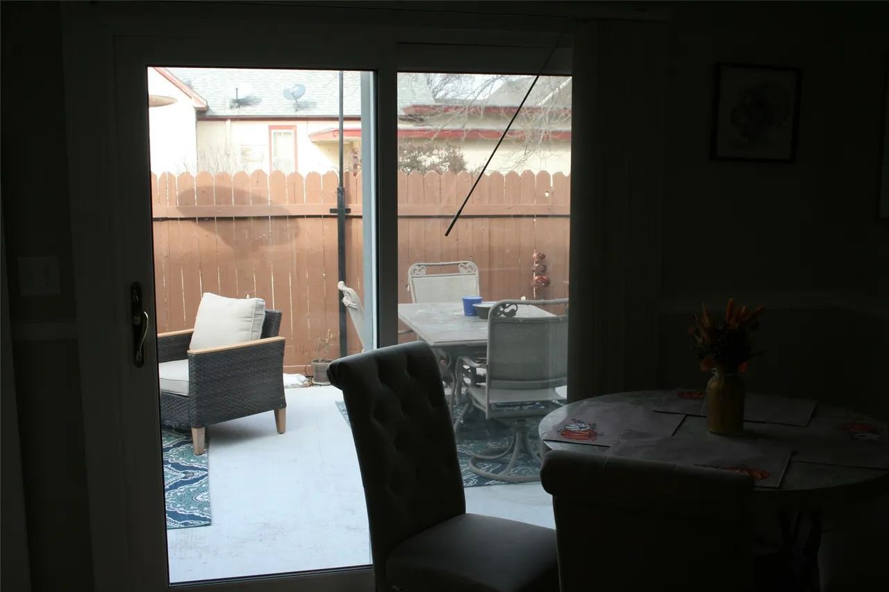 A view from inside looking out through a glass door at a patio with a chair, a dining table, and a wooden fence.