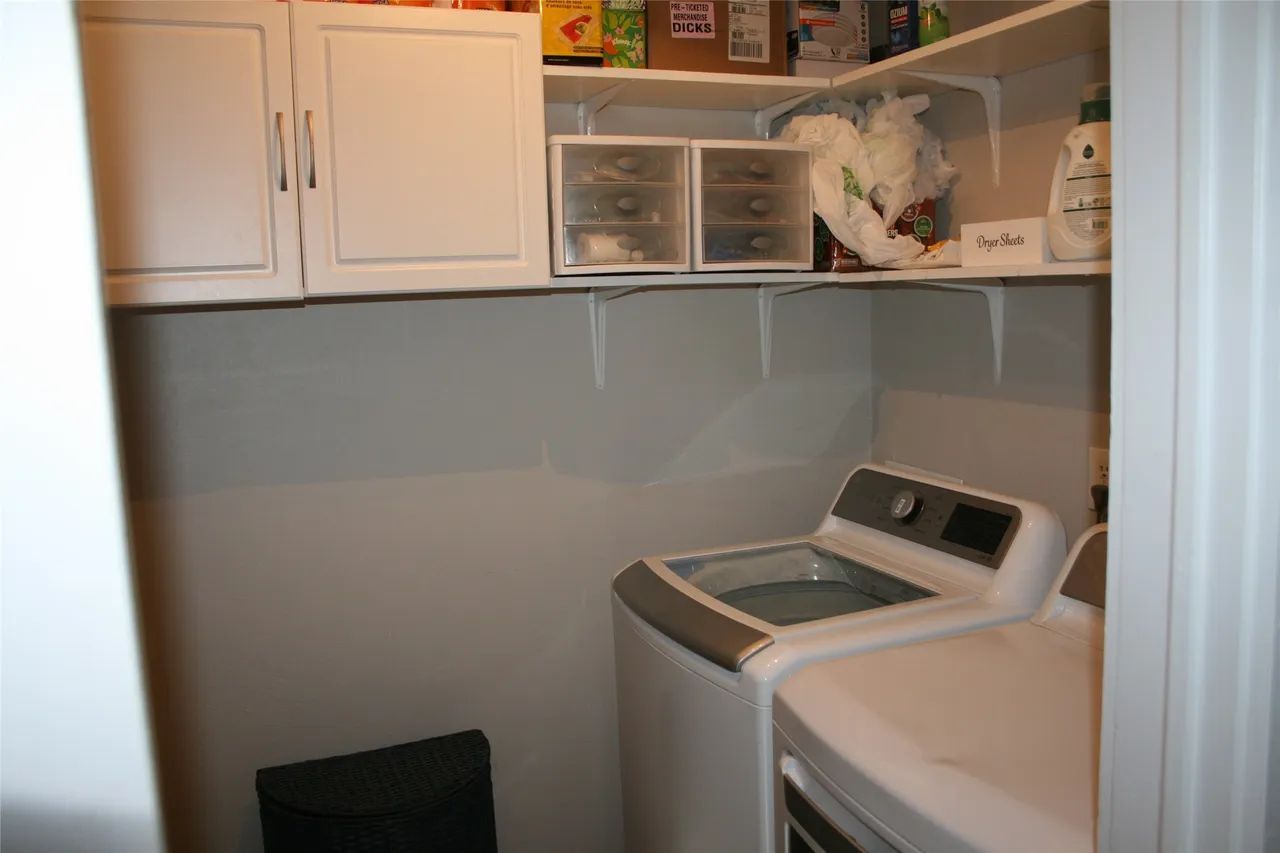 Laundry room with white cabinets, open shelving holding storage bins and supplies, and a top-loading washer and dryer.