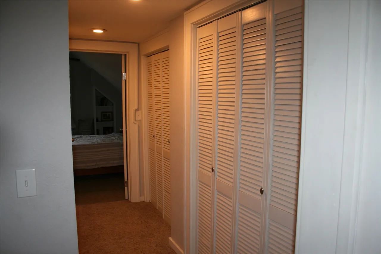 A hallway with light carpet, beige walls, and two sets of white louvered bifold closet doors leading to a dark room.