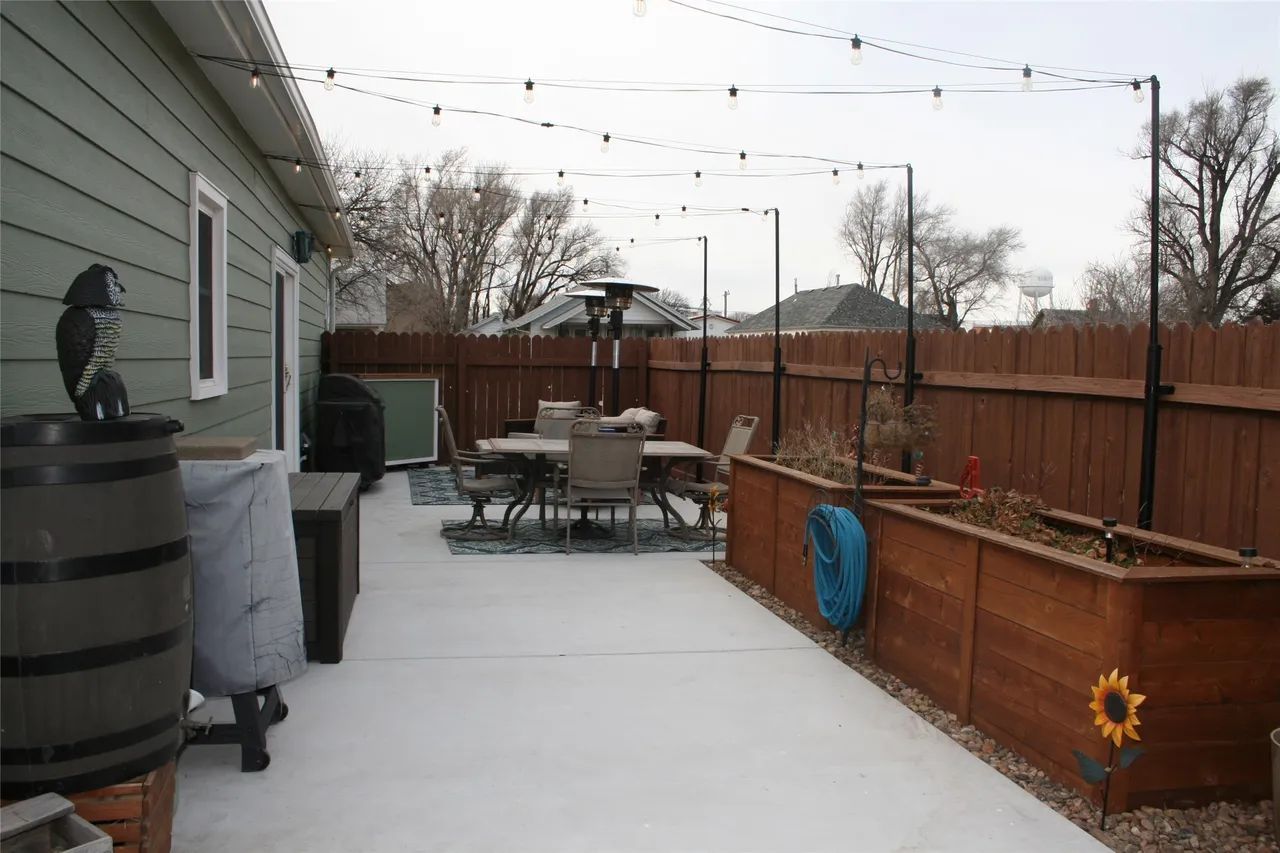 Patio area with a dining set, raised wooden garden beds, string lights, and a wooden fence on a light-colored concrete slab.