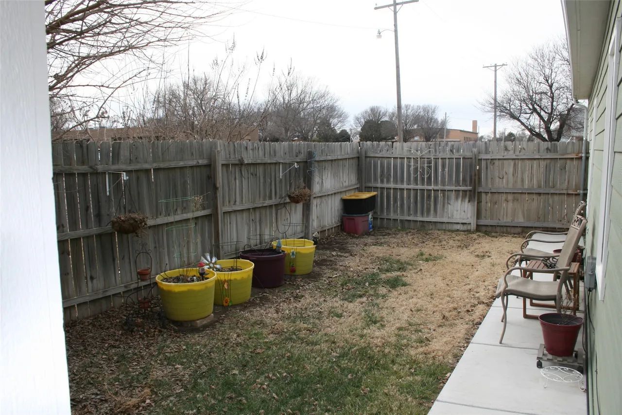 A small backyard with a wooden fence, several yellow and dark pots, and two chairs on a concrete patio.