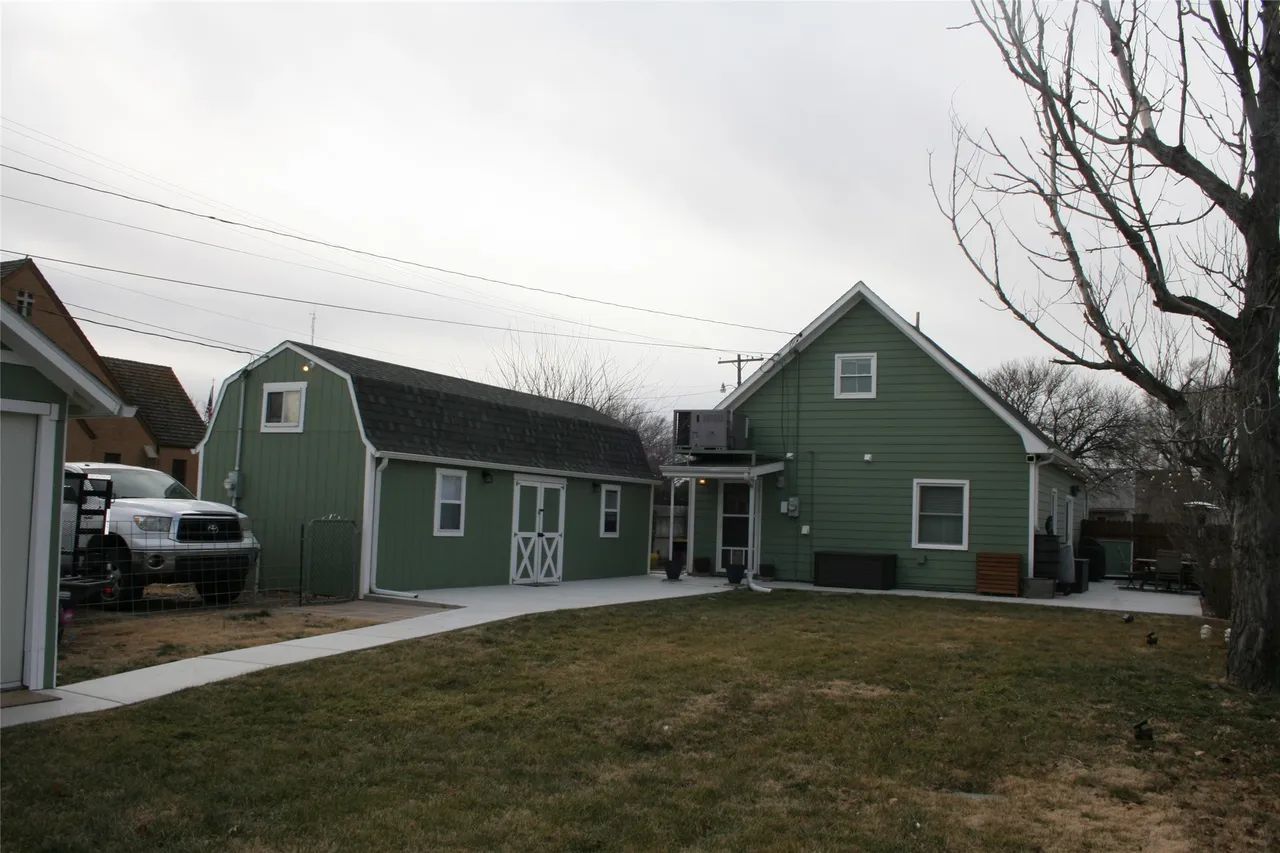 A green house and adjacent gambrel-roofed shed sit on a property with a concrete walkway and a grass yard.