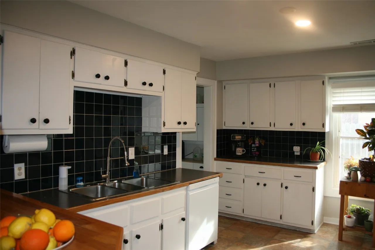 A kitchen with white cabinets, dark tile backsplashes, wood countertops, a stainless steel sink, and a window.