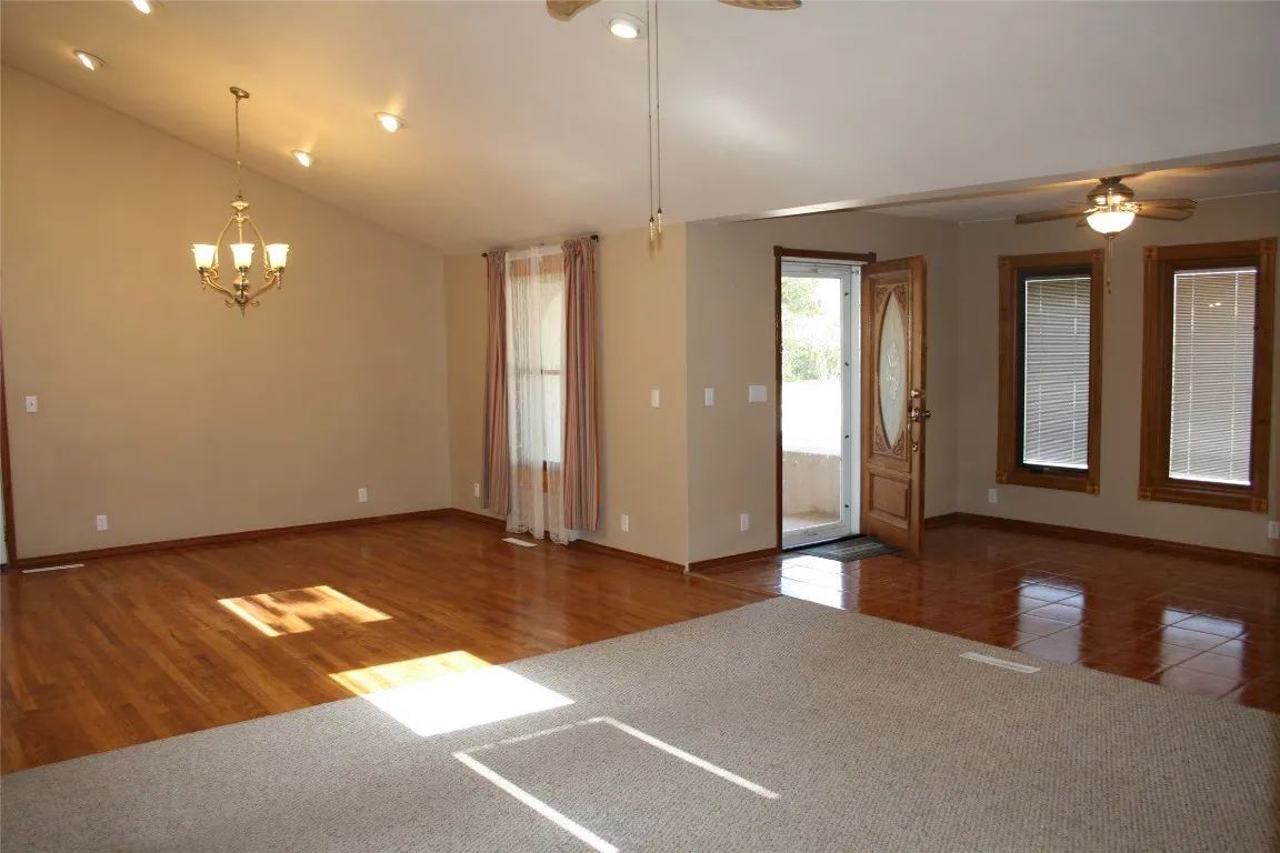 A spacious room with hardwood floors, a gray carpeted area, high ceilings, a chandelier, and a door leading outside.