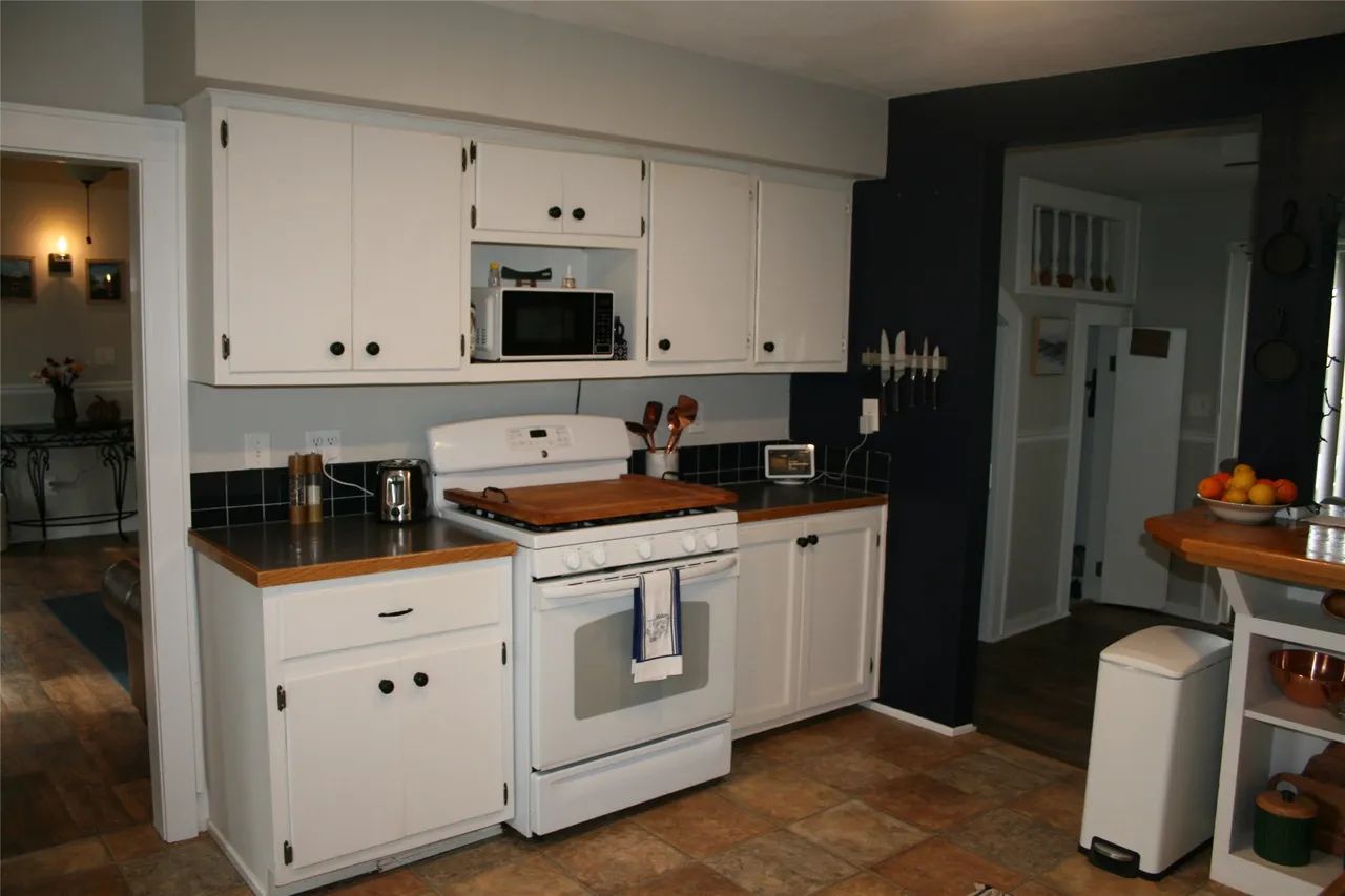 A white-cabinet kitchen with a stove, microwave, and dark walls, opening into a tiled floor area with a trash can.