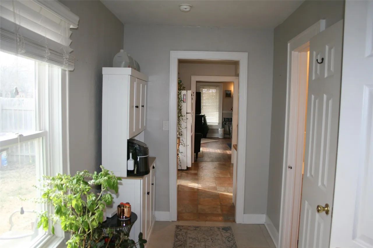 A hallway with light gray walls, a window with blinds, a white cabinet, a potted plant, and a doorway to a dining room.