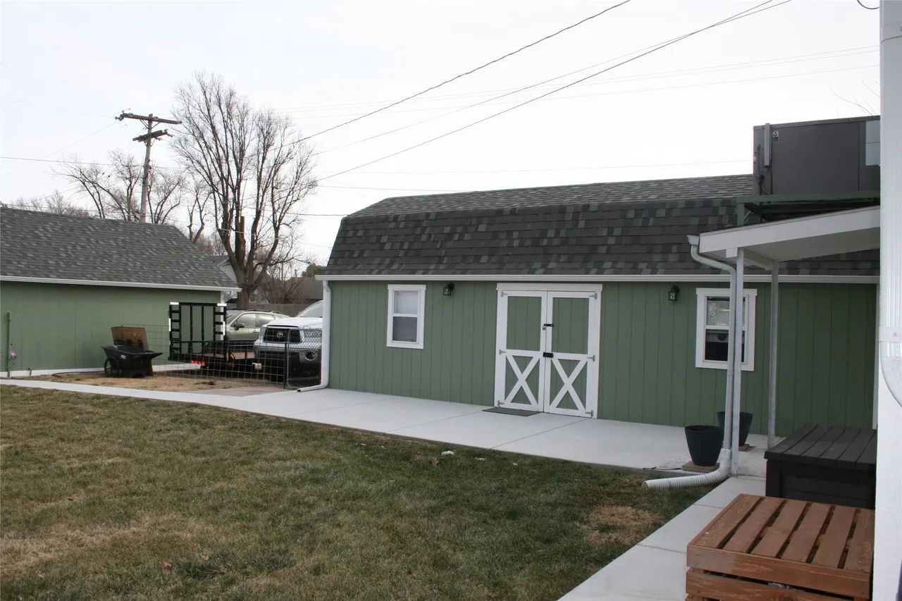 A green shed with white trim and double doors sits beside a house, bordering a concrete walkway and a grassy lawn.