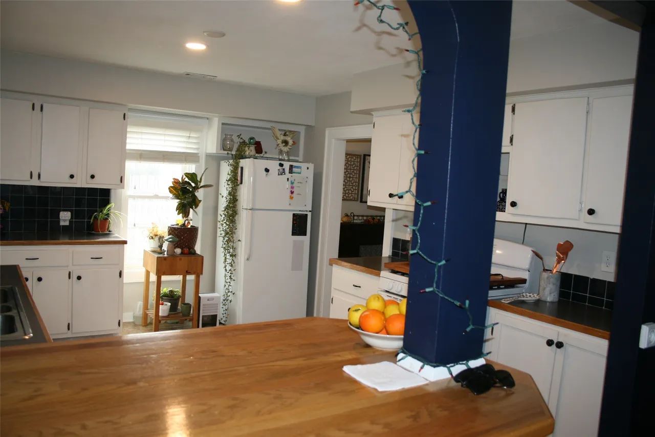 A view of a kitchen with white cabinets, a dark blue structural pillar, and a wooden island countertop with a bowl of fruit.