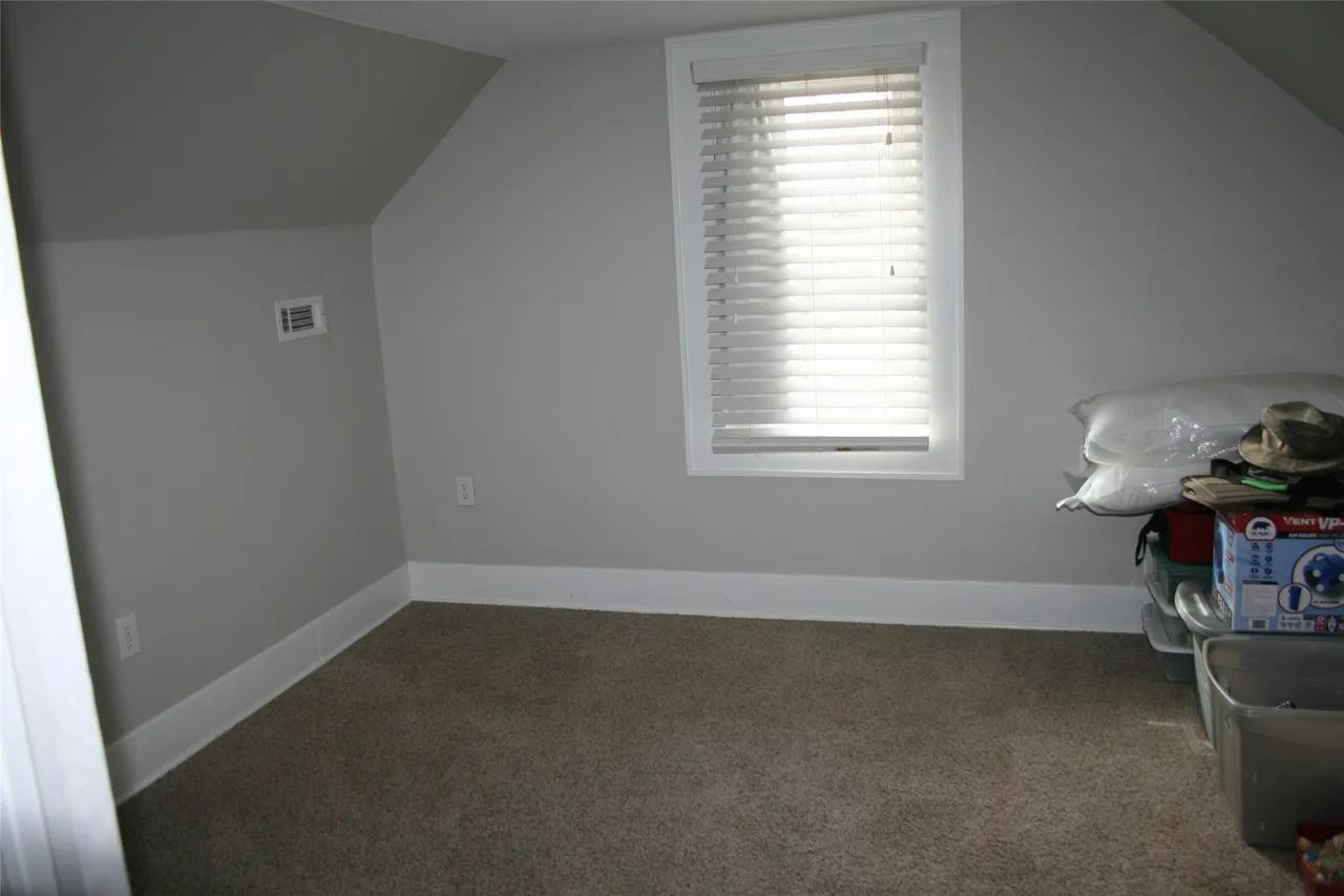 A sparsely furnished room with light gray walls, carpeted flooring, a window with closed blinds, and storage bins.