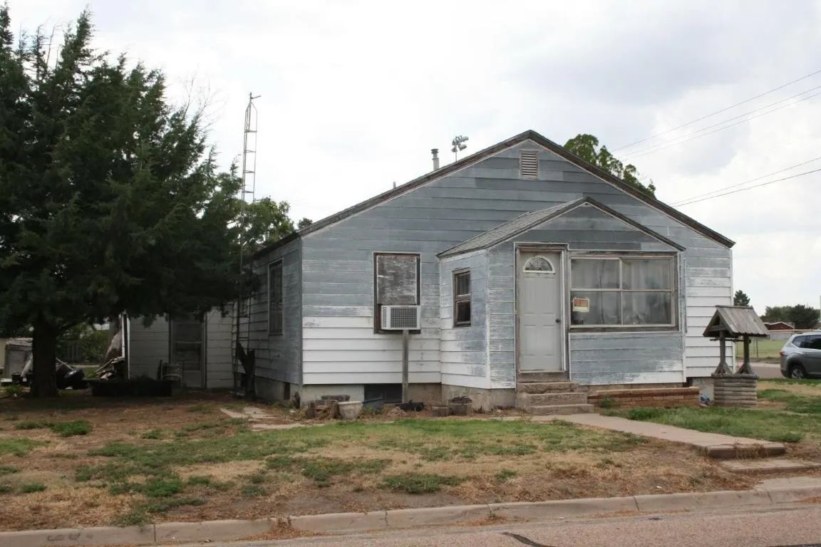 A small, one-story house with weathered grey and white siding, an air conditioning unit, and a tree in the front yard.